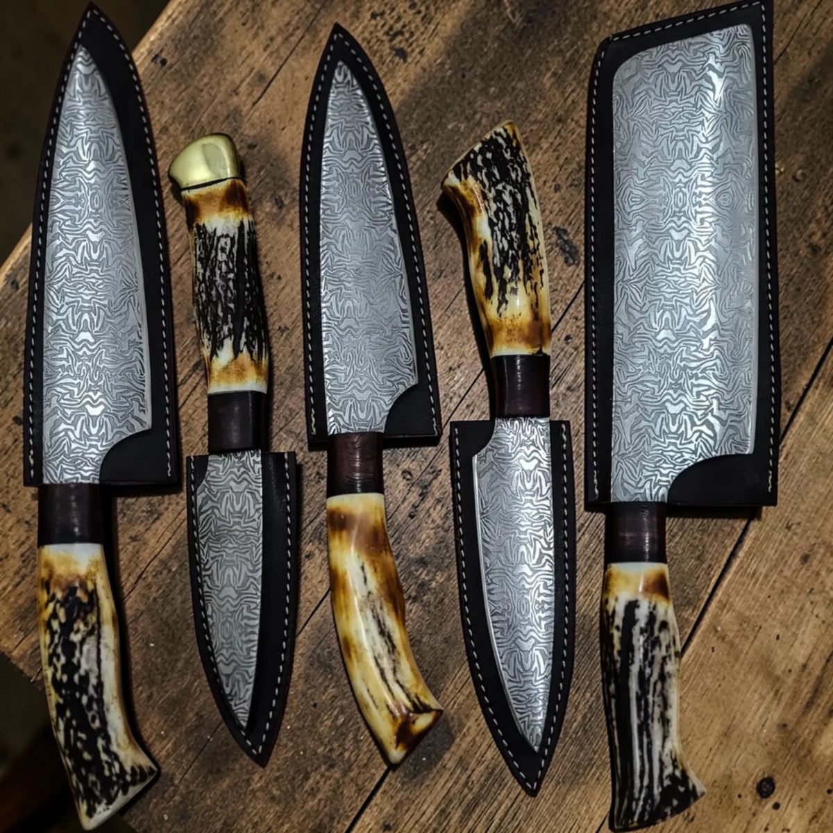Handmade Damascus culinary knife set featuring antler handles inside fitted leather sheaths on a rustic surface