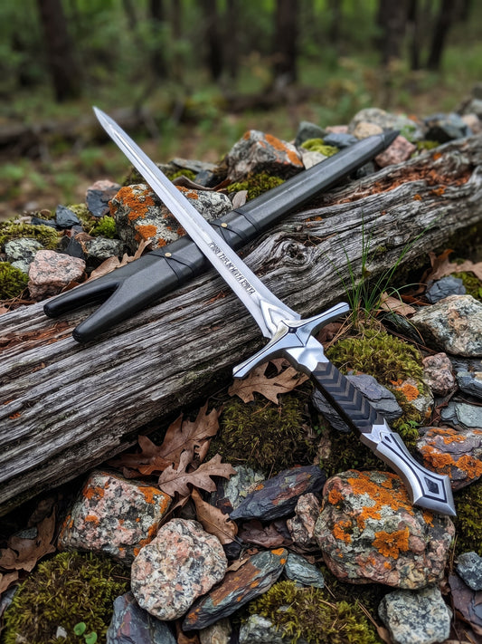 Stainless steel fantasy sword with textured rubber wrapped handle and decorative scabbard with polished steel chape displayed on woodland rocks