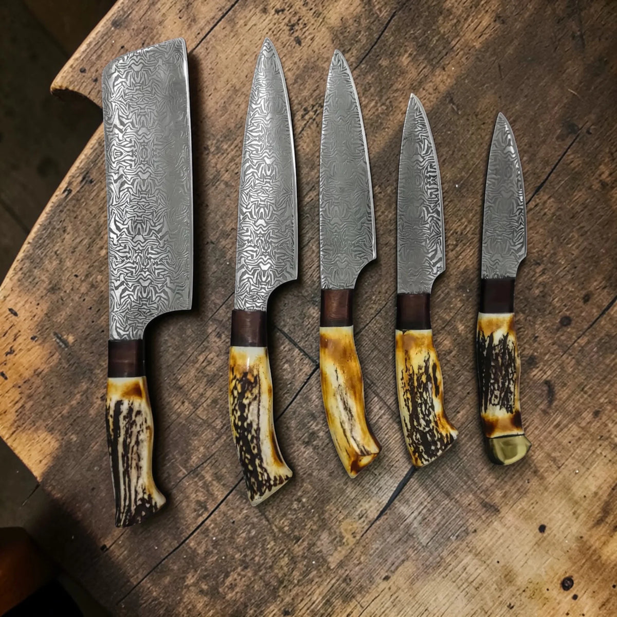 5 piece Damascus steel knife set with unique antler and brass handles laid out on a wooden background