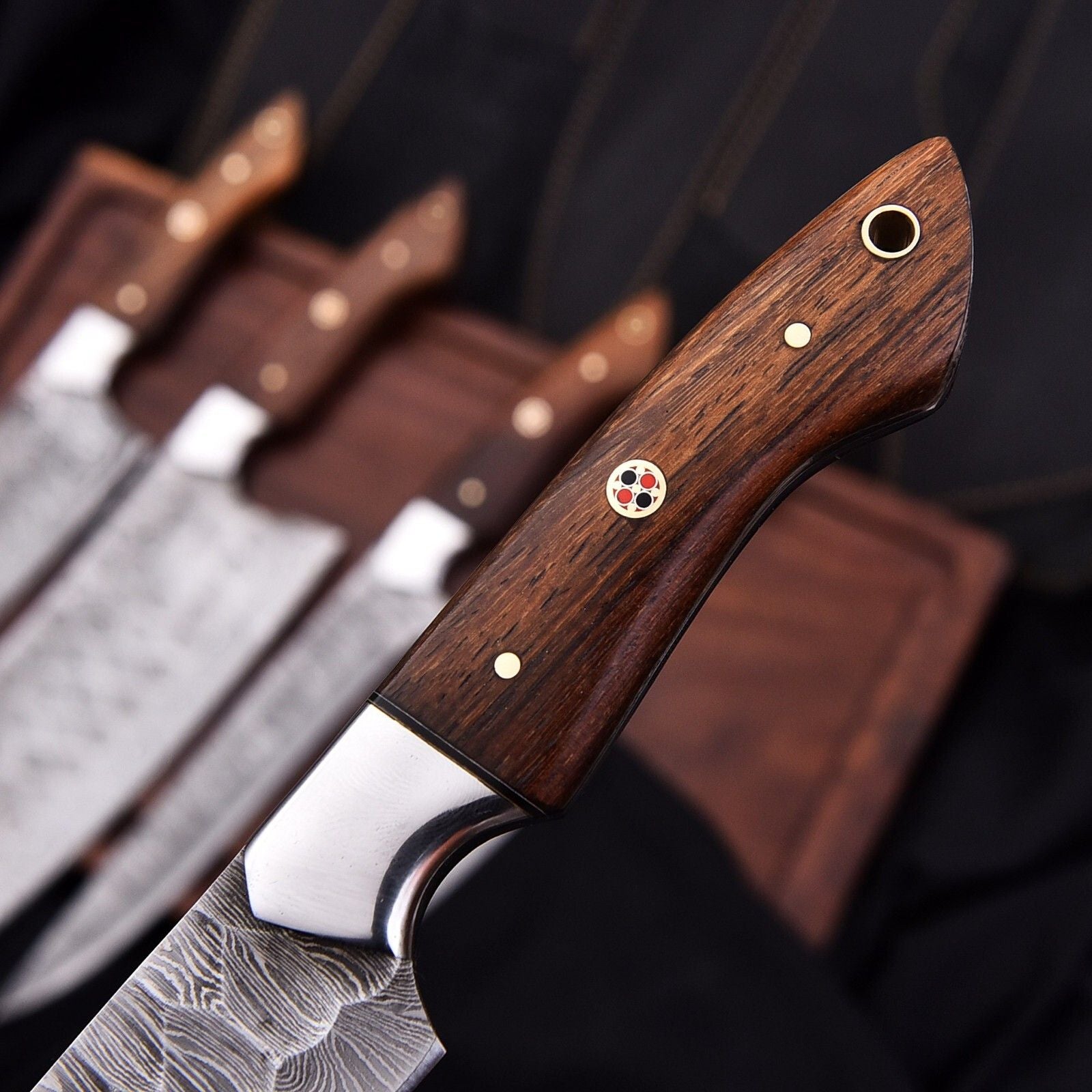 Close up detail of a Damascus steel kitchen knife handle showcasing artisan rosewood, stainless steel bolster, and decorative mosaic pin.