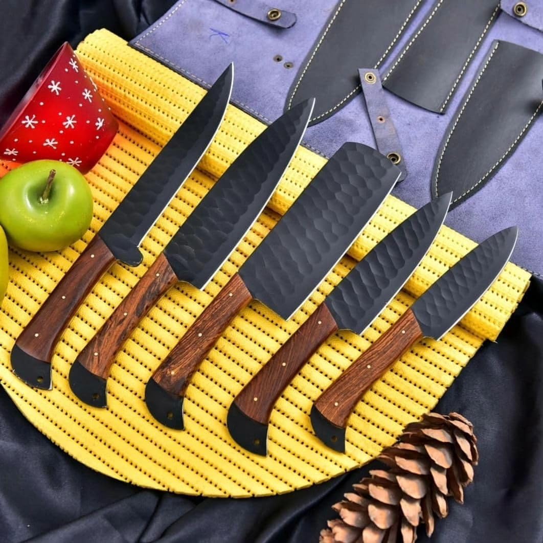 Five black hammered kitchen knives with rosewood handles displayed on a yellow woven mat beside a green apple and pine cone.