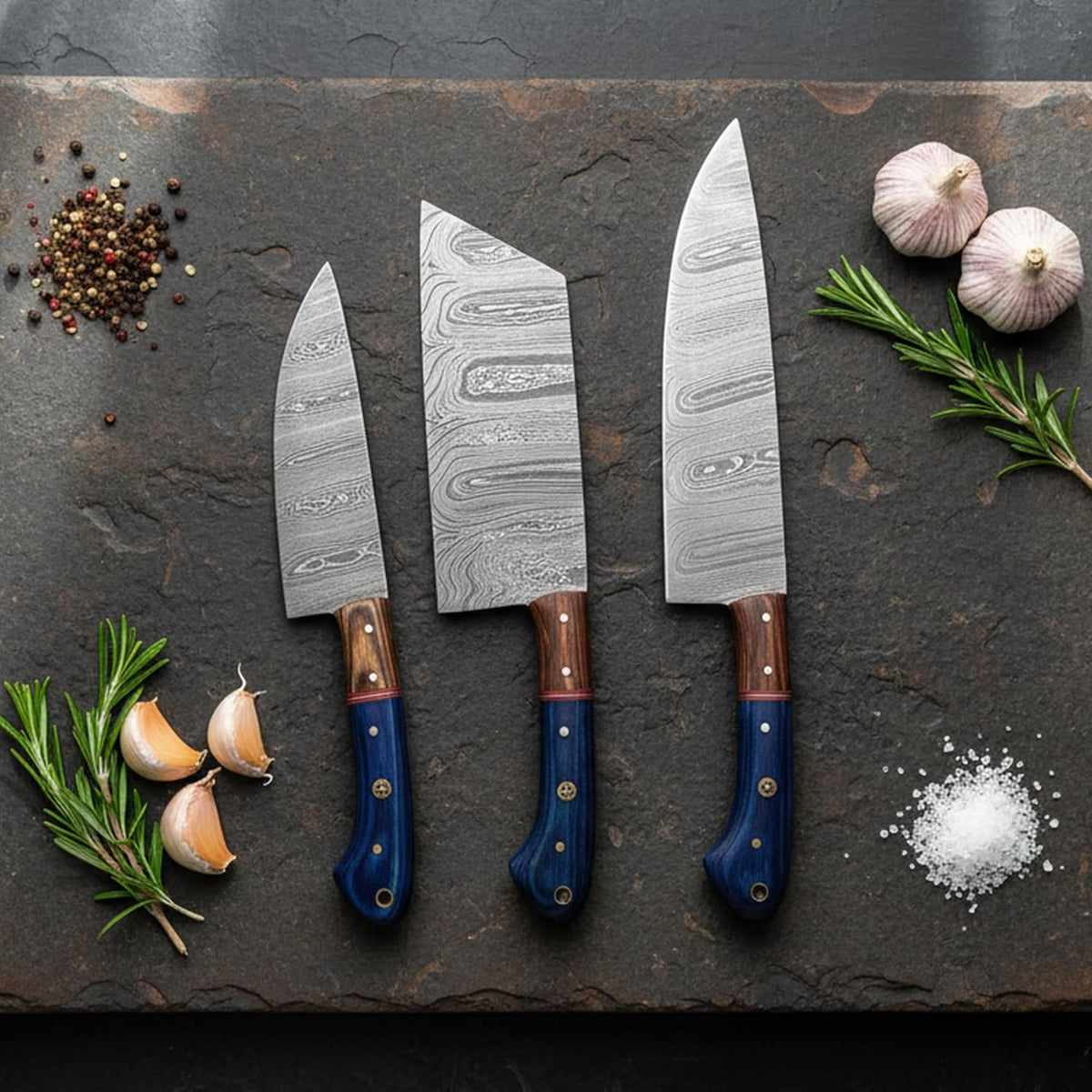 Three Damascus kitchen knives with blue wood handles on slate with garlic and salt