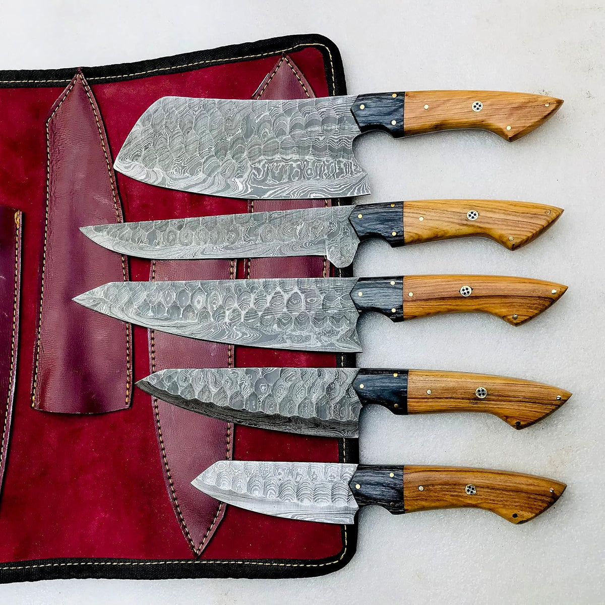Damascus chef knife set of five laid across an open burgundy leather knife roll