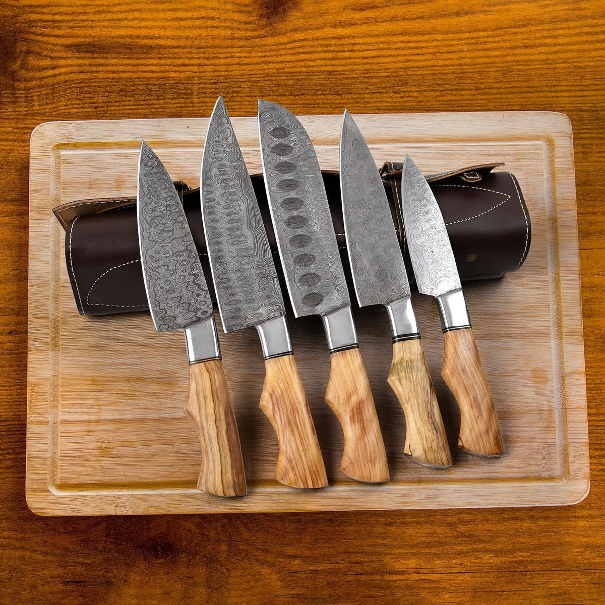 Damascus knife set with pakka wood handles fanned on a wood cutting board next to its leather roll case
