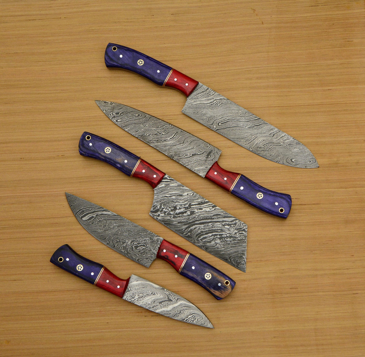 Five Damascus kitchen knives with purple and red pakkawood handles on light wood