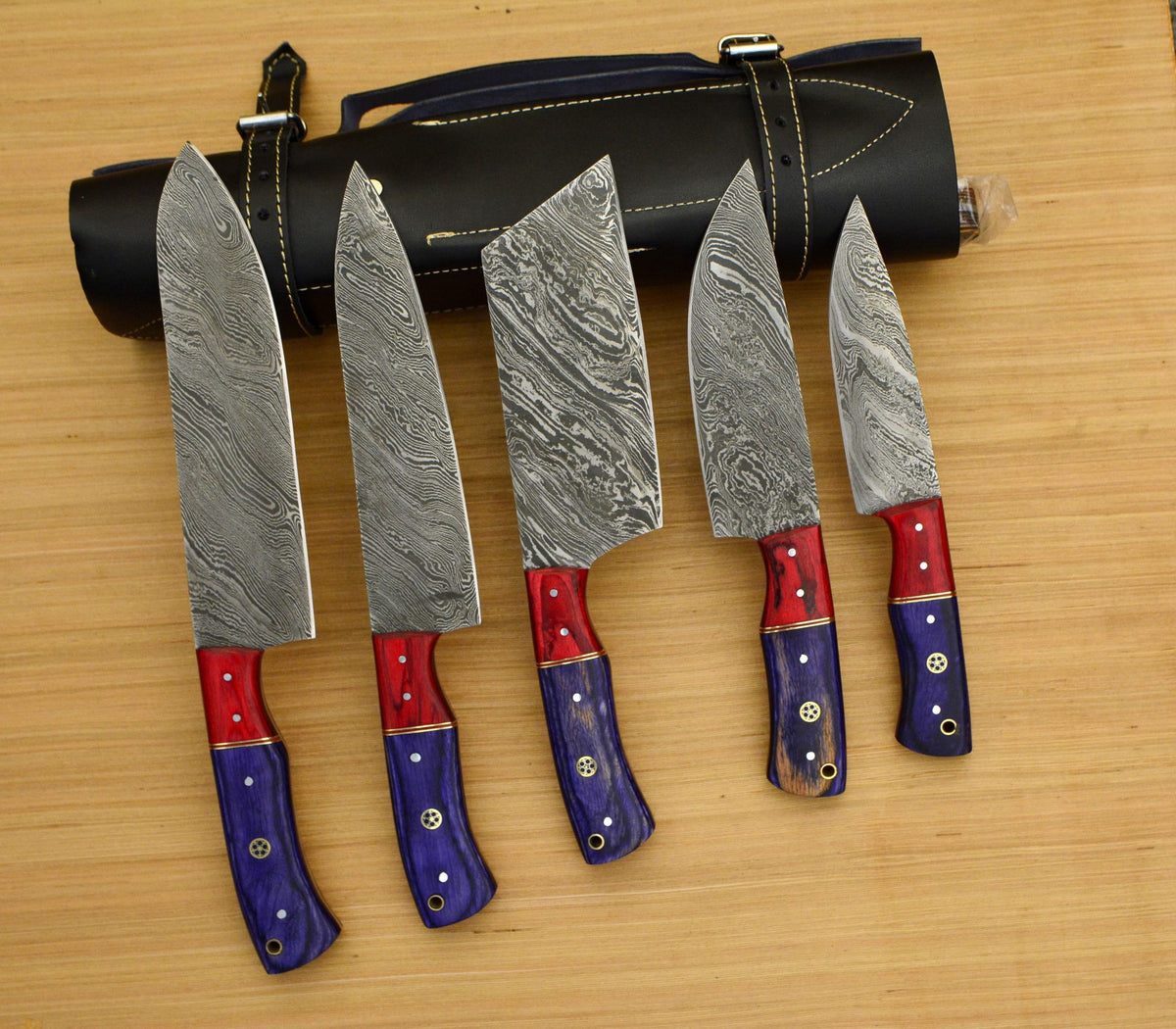 Five Damascus knives with purple and red handles in front of a black leather roll