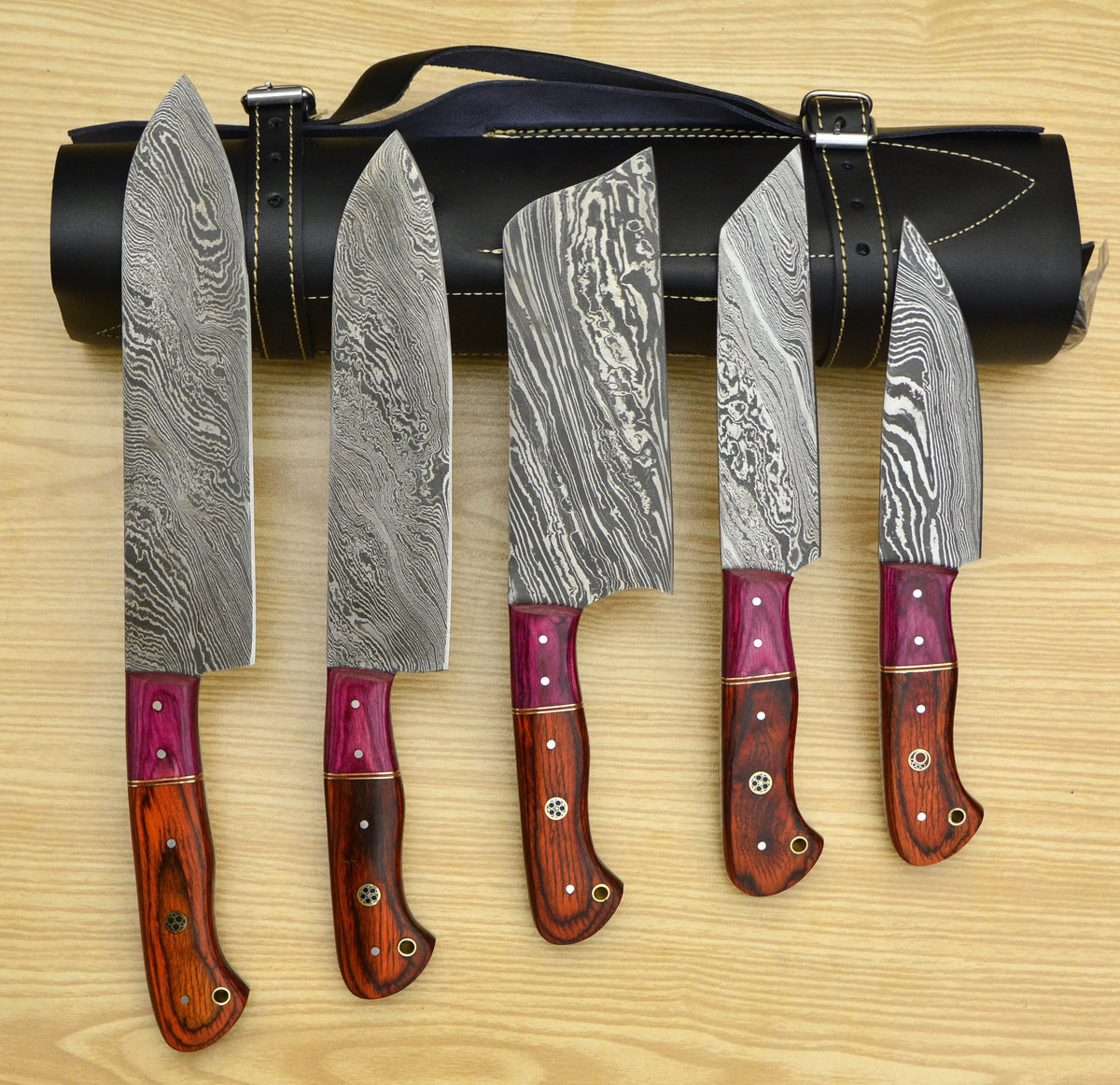 Damascus chef knife set with red pakka wood handles beside black leather roll
