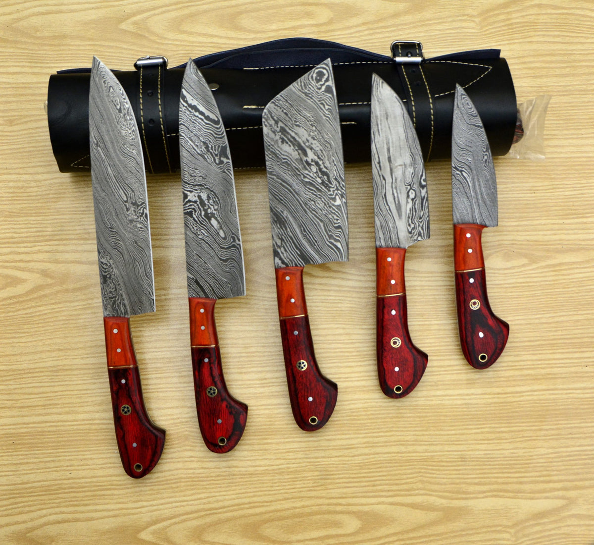 5 pcs Damascus steel chef knives with red wood handles arranged upright next to a rolled leather bag on a wooden surface