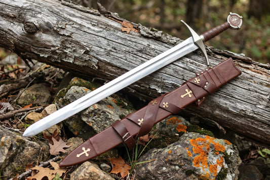 Decorative sword with grooved blade beside a brown leather sheath with gold tone crosses and a buckled strap on rocks