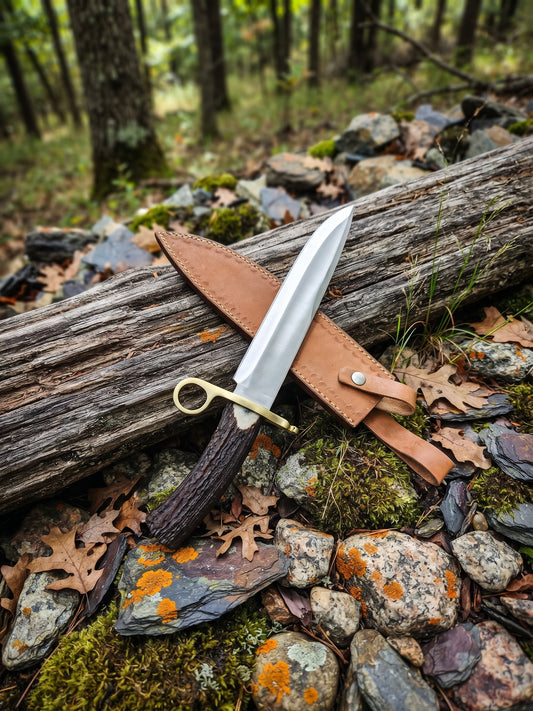 Crown antler Bowie knife with stainless steel blade and leather sheath displayed on a woodland log