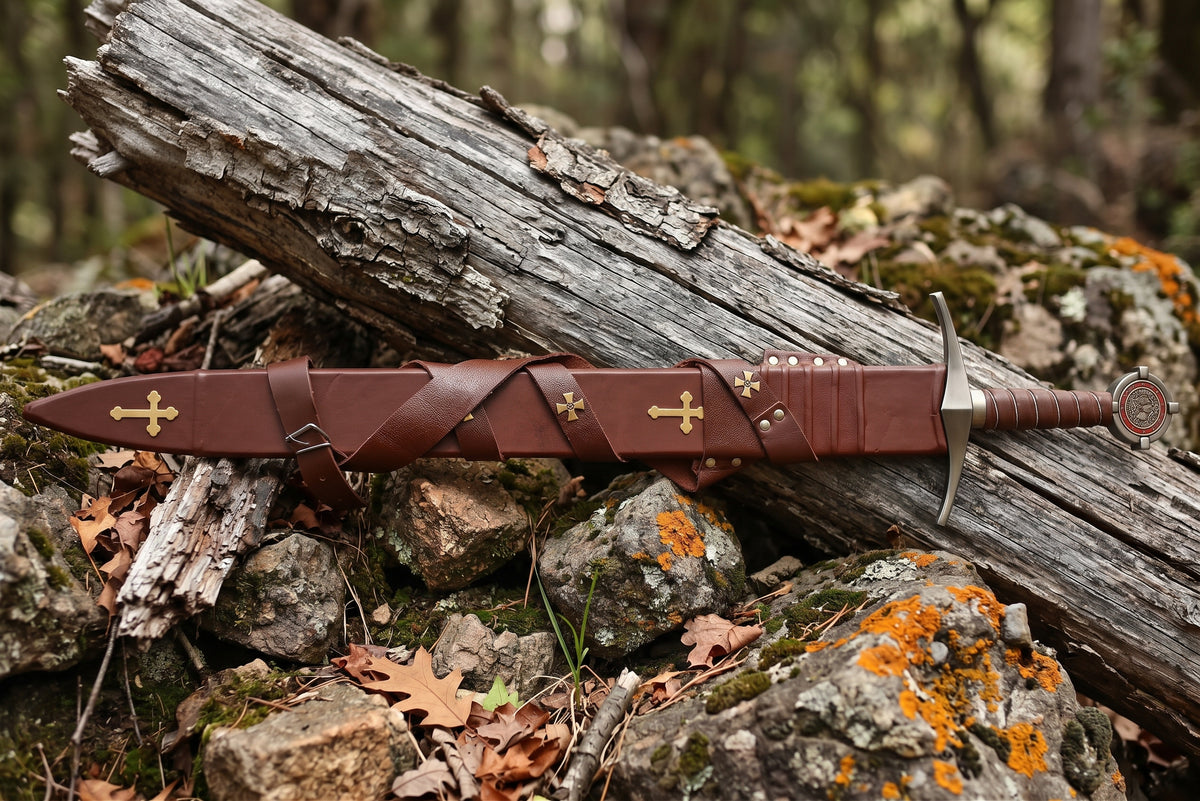 Brown leather sheath with gold tone cross plates and buckled strap on a log with a decorative sword hilt at the end
