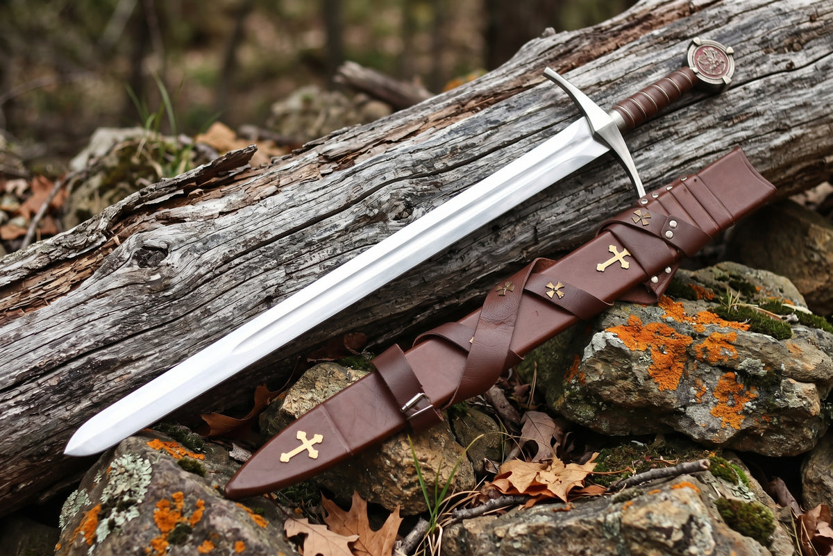 Decorative sword lying beside a brown leather sheath with gold tone cross plates and a criss cross strap on mossy rocks