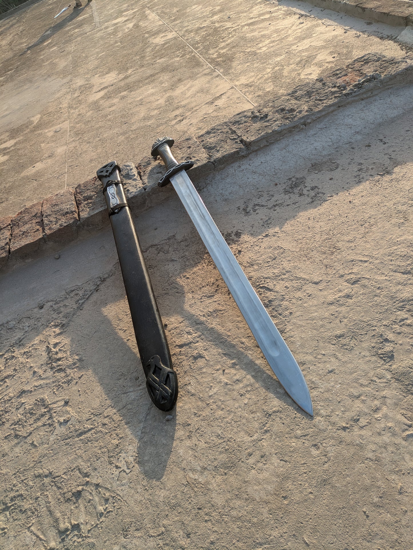 Handmade Fantasy Sword with polished blade resting beside black wooden scabbard outdoors