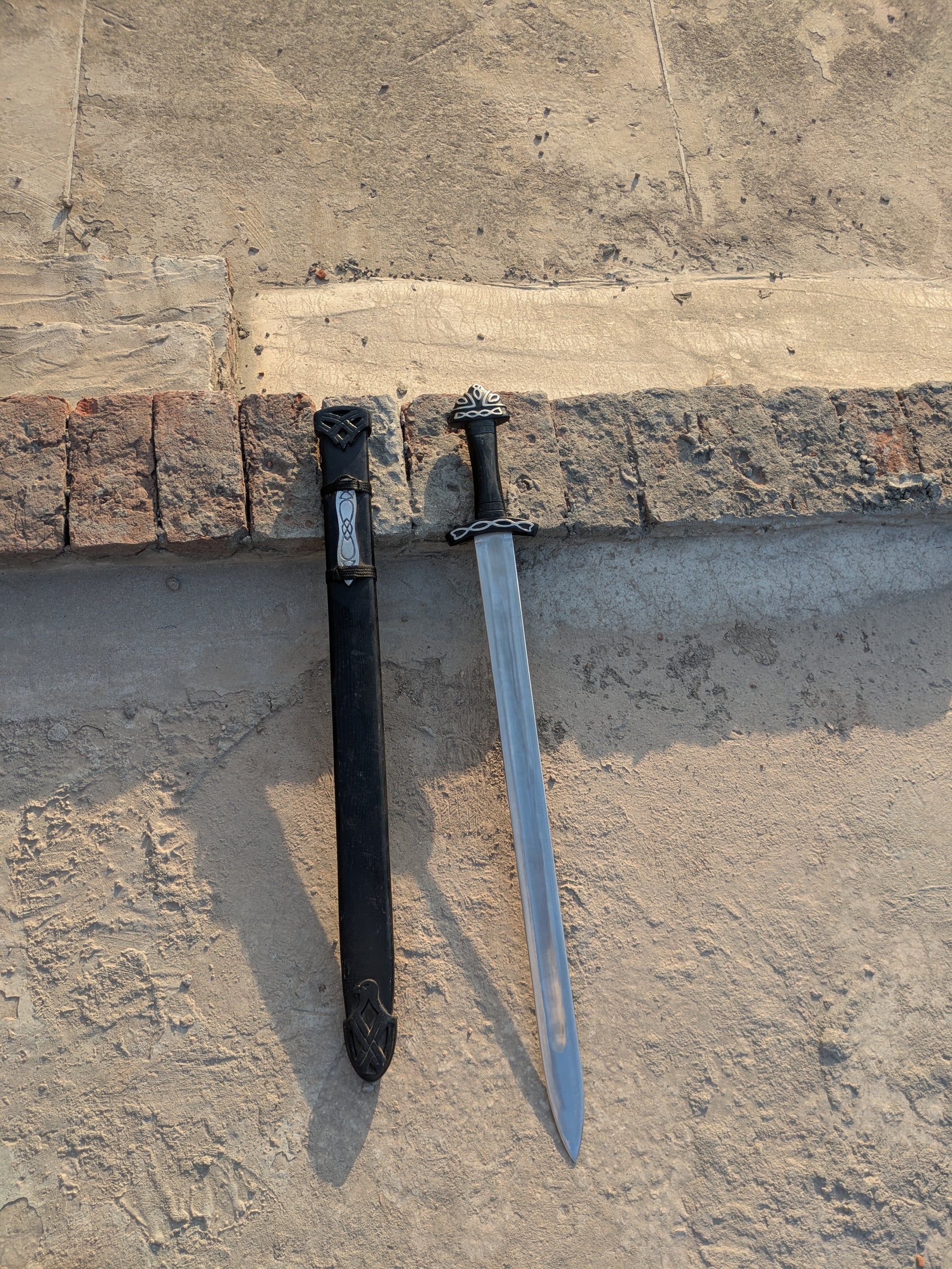 Handmade Fantasy Sword full length shown with black wooden scabbard against brick wall