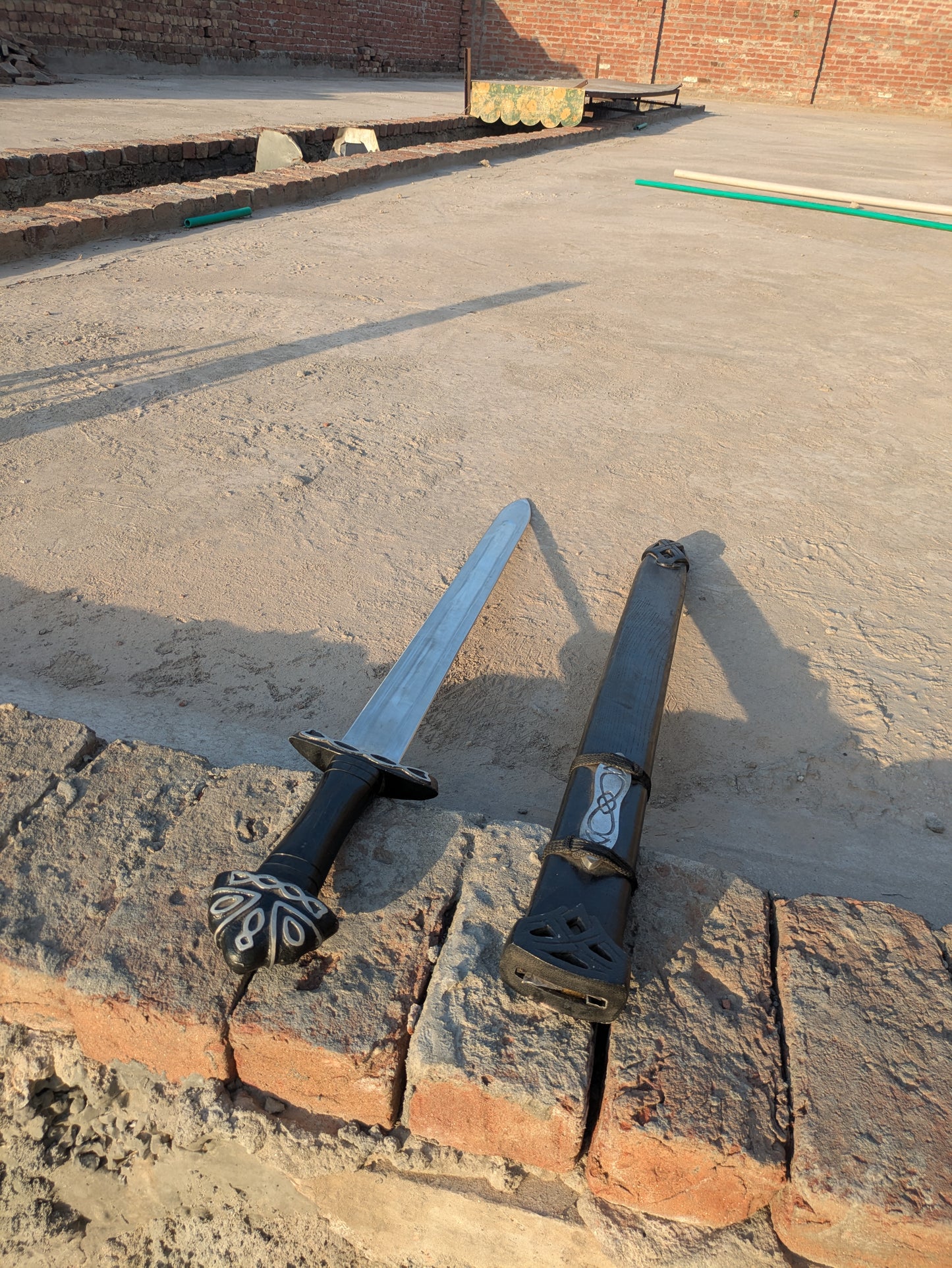 Handmade Fantasy Sword with carved pommel resting beside black wooden scabbard outdoors