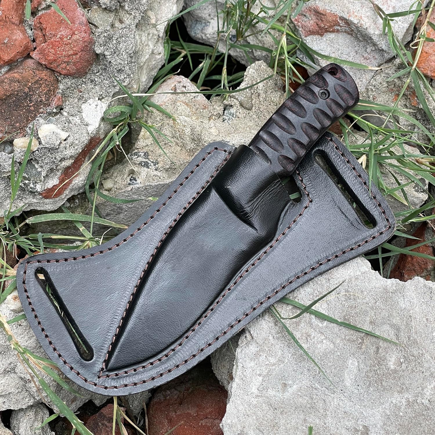 A hand grips the best skinning knife in front of a dark, rustic stone archway leading into a moody forest. The knife features a black powder-coated drop-point blade and a dark, deeply grooved ergonomic handle. Behind the knife rests its heavy-duty black leather pancake sheath with thick perimeter stitching. This atmospheric outdoor setting perfectly highlights the rugged nature of this premium hunting tool.