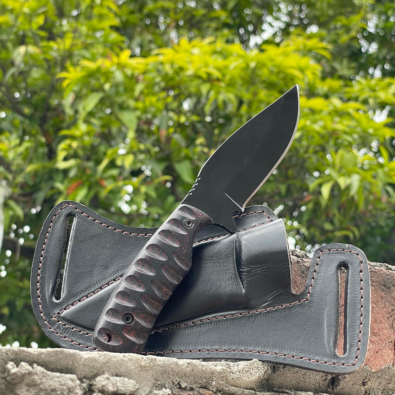 Outdoor shot of the best skinning knife resting against its black leather sheath on a textured brick ledge. The fixed blade features a black powder-coated carbon steel blade with a drop point and a dark Micarta handle with deep scalloped grooves for a secure grip. The rugged black leather pancake sheath shows prominent stitching and belt loops. A softly blurred background of lush green foliage highlights this hunting tool's outdoor readiness.