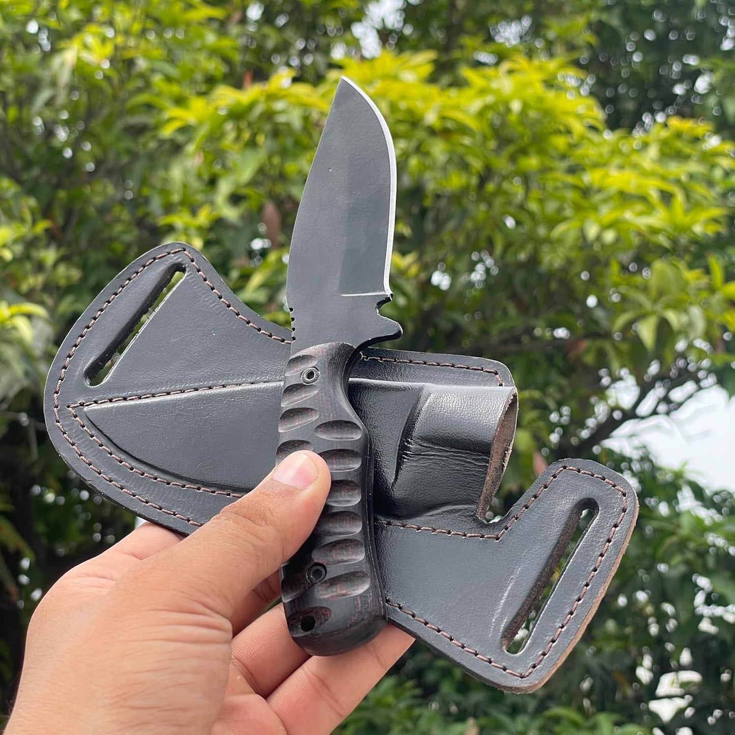 A hand securely grips the best skinning knife outdoors, showcasing its dark, grooved Micarta handle and black powder-coated carbon steel blade. A heavy-duty black leather pancake sheath, featuring thick stitching and horizontal belt loops, rests behind the knife. The background is a soft blur of green foliage, emphasizing the knife's readiness for hunting and field dressing.