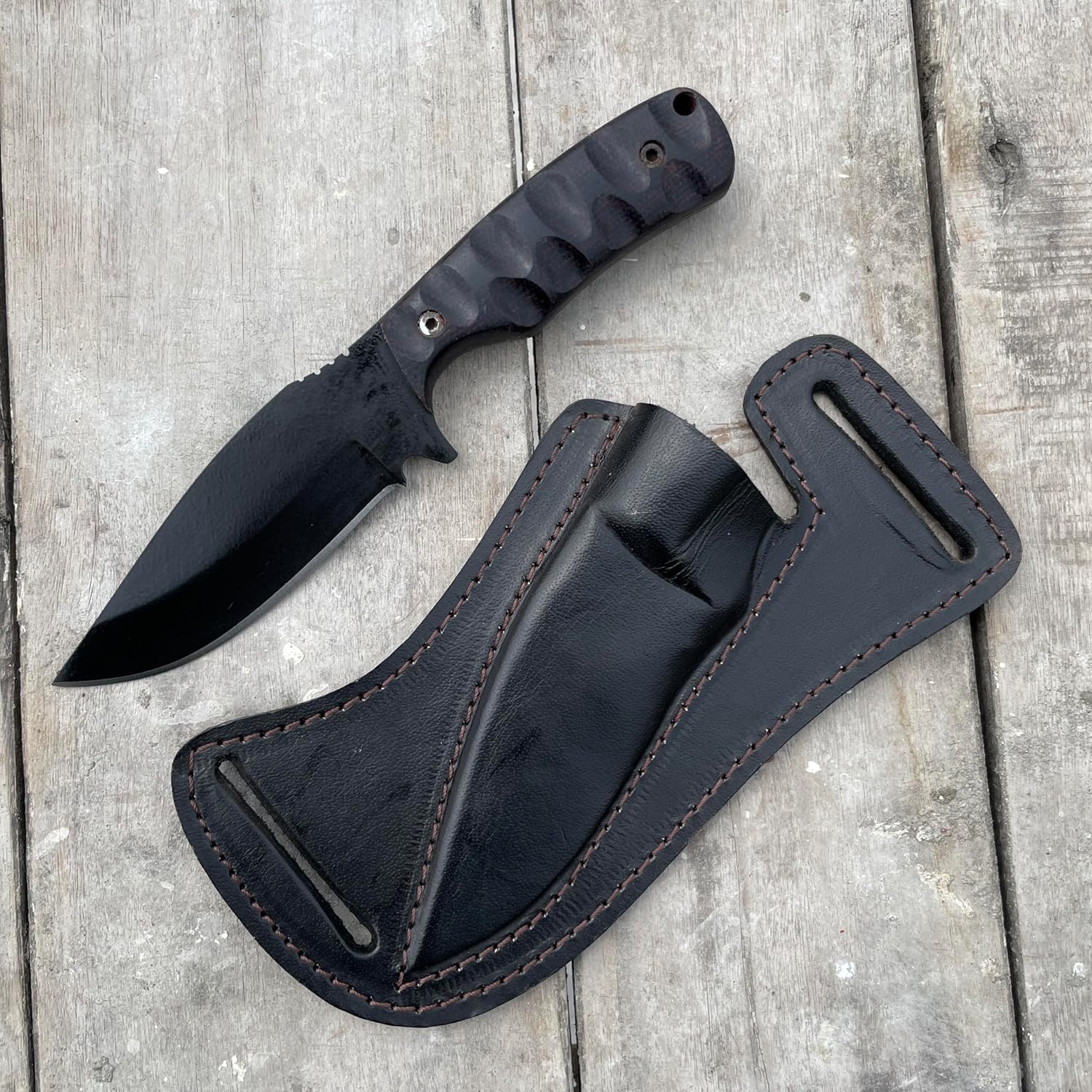 Regarded as the best skinning knife, this sleek fixed-blade hunting knife rests on weathered wooden planks beside its matching black leather pancake sheath. The knife features a sharp, black powder-coated drop-point blade and a dark, heavily grooved Micarta handle with a lanyard hole. The rugged sheath showcases prominent brown stitching and dual belt slots designed for secure horizontal carry, making it a perfect tool for outdoor excursions and field dressing.