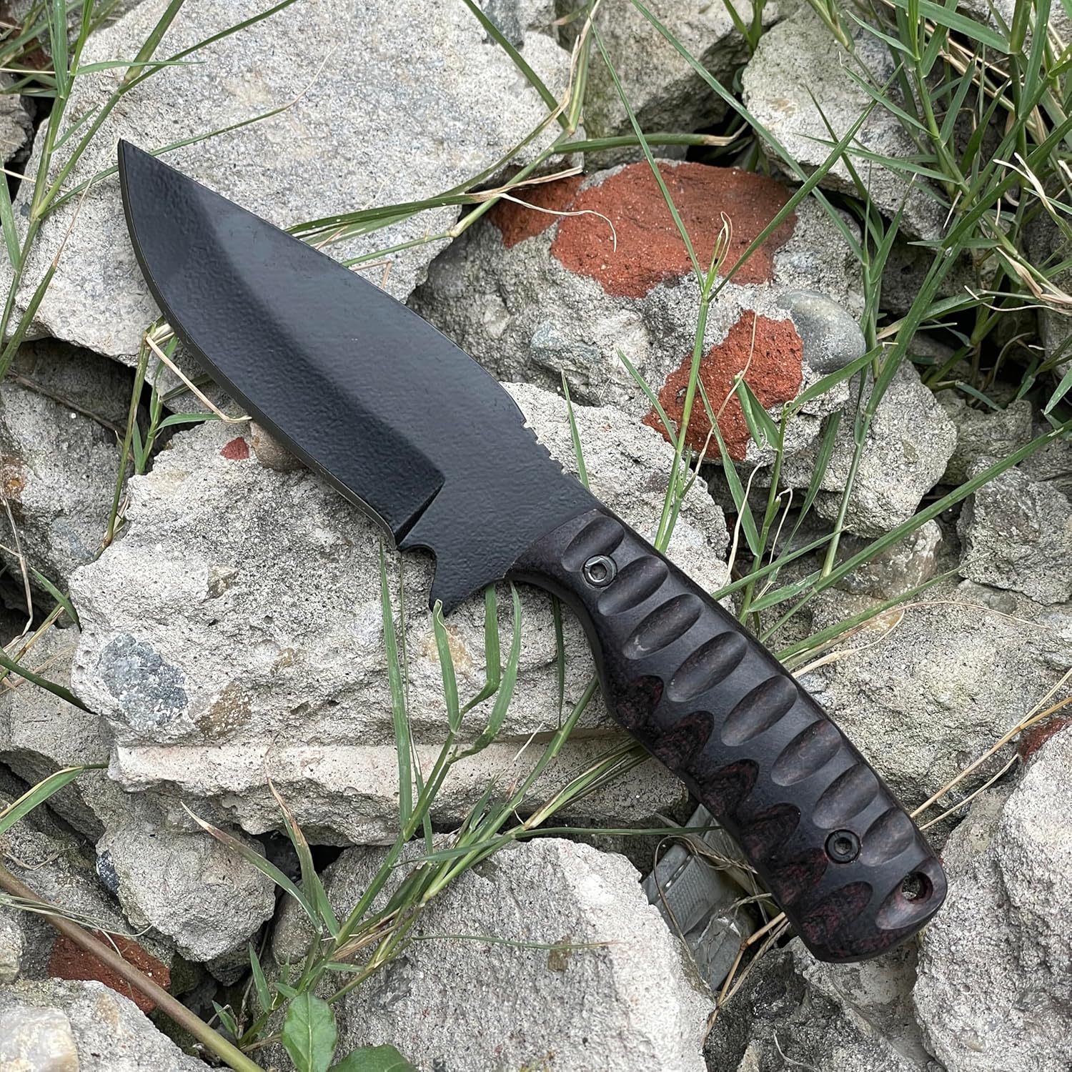 Considered the best skinning knife for outdoor use, this rugged fixed-blade knife rests on a bed of broken grey concrete, red brick fragments, and green grass. The tactical knife features a matte black powder-coated drop-point blade and a dark, deeply grooved ergonomic handle secured with two pins. A lanyard hole sits at the base. The natural outdoor setting highlights the knife's durable construction and readiness for demanding hunting and field dressing tasks.