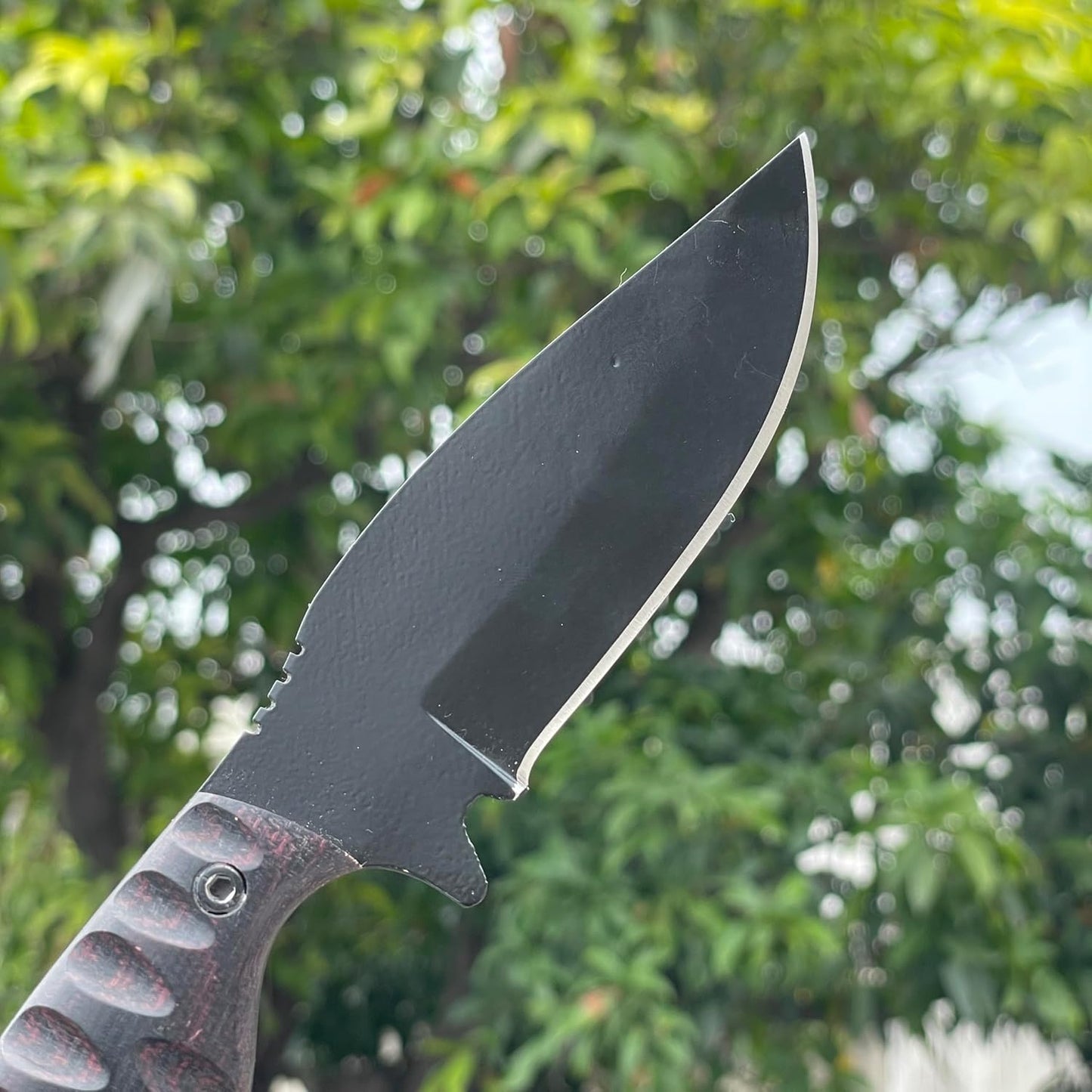 Close-up of the best skinning knife held against a softly blurred background of lush green foliage. This premium hunting knife features a matte black powder-coated drop-point blade with a bright, razor-sharp edge and jimping on the spine for added control. The dark Micarta handle showcases deep, scalloped grooves and a visible metal pin, highlighting its ergonomic design and secure grip for precise field dressing.