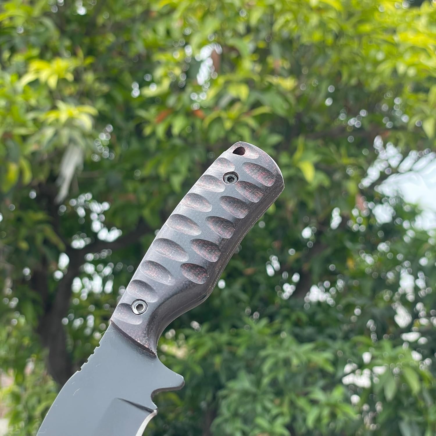 Close-up focusing on the ergonomic handle of the best skinning knife, held against a softly blurred background of green foliage. The dark Micarta handle features deep, scalloped grooves for a secure grip, secured by two metal screws, and includes a lanyard hole at the base. A portion of the matte black powder-coated blade with a prominent finger guard is visible, highlighting the rugged and reliable construction of this premium outdoor hunting tool.