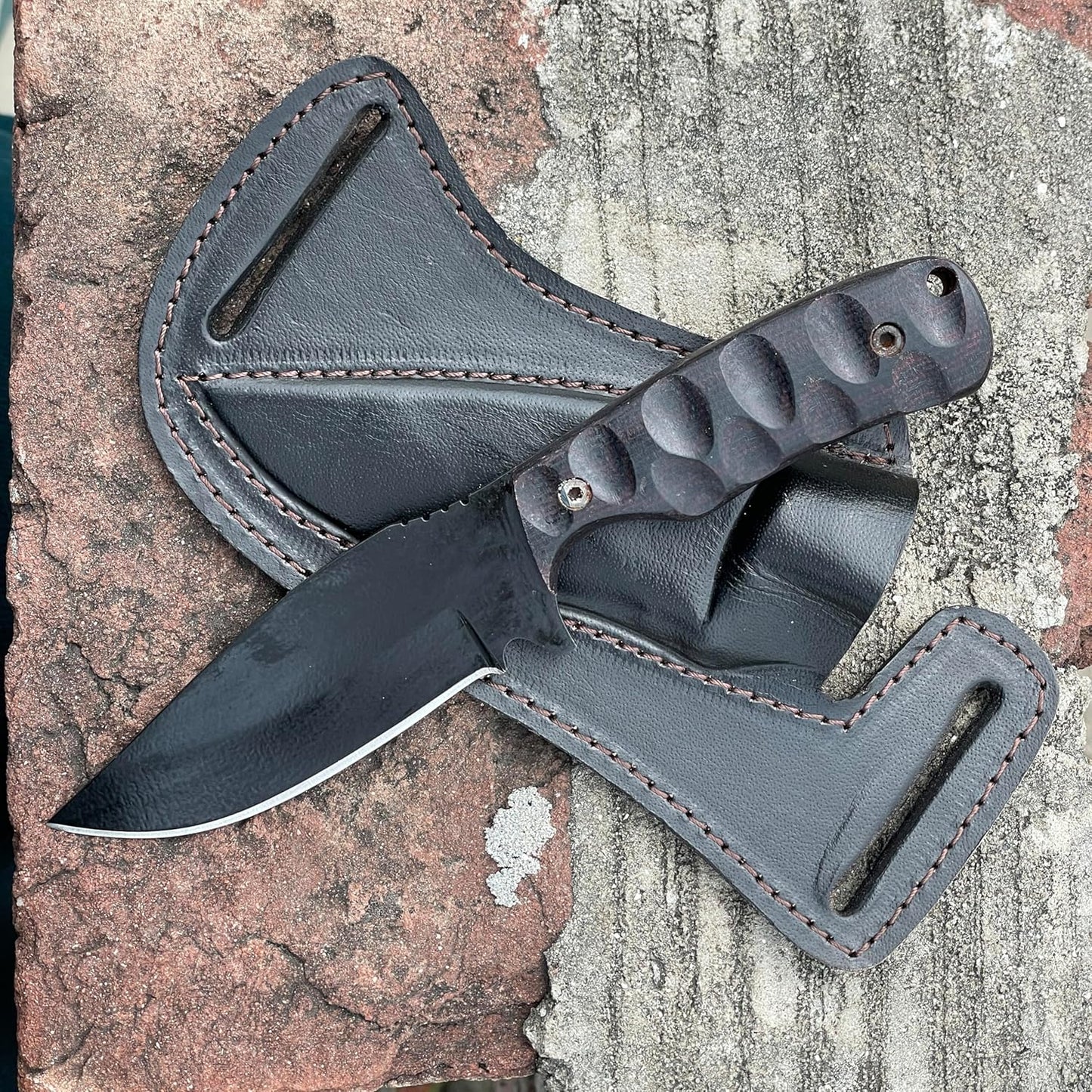 Resting on weathered bricks, the best skinning knife features a black powder-coated drop-point blade with a sharp silver edge. Its dark Micarta handle has deep scalloped grooves for a secure grip, fastened with metal pins. The knife lies across its custom black leather pancake sheath, which highlights thick brown stitching and dual belt loop slots for horizontal carry. This rugged setup showcases a premium tool ready for hunting and field dressing.