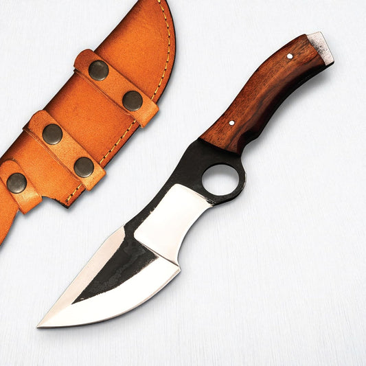 A hunting tracker knife resting diagonally on a white textured surface alongside a light brown leather sheath. The knife features a polished wooden handle secured with two silver pins and a flat, exposed metal pommel. The distinct two-toned blade has a dark, forge-textured upper section featuring a large circular finger hole near the handle, contrasting with the highly polished silver, multi-faceted cutting edges. The accompanying tan leather sheath displays visible yellow stitching and two retaining straps