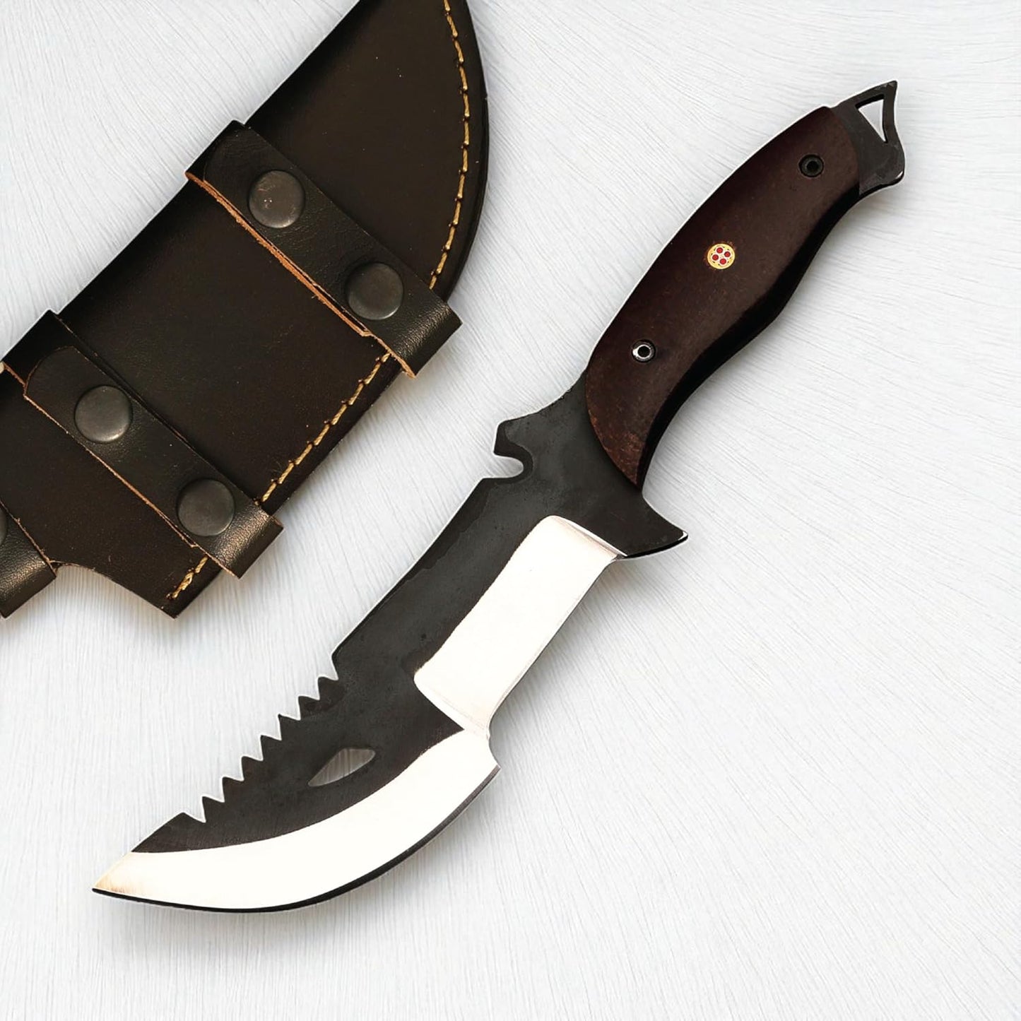 A heavy-duty survival tracker knife resting on a white textured surface next to a dark brown leather sheath with snap closures. The knife features a dark wood handle secured with a decorative mosaic pin, a full tang with an exposed lanyard loop at the pommel, and a distinct two-toned blade. The blade has a matte black upper section with saw teeth along the spine and a teardrop-shaped cutout, contrasting with the polished silver, compound cutting edge designed for both chopping and slicing.
