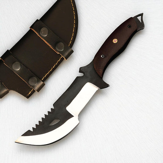 A heavy-duty survival tracker knife resting on a white textured surface next to a dark brown leather sheath with snap closures. The knife features a dark wood handle secured with a decorative mosaic pin, a full tang with an exposed lanyard loop at the pommel, and a distinct two-toned blade. The blade has a matte black upper section with saw teeth along the spine and a teardrop-shaped cutout, contrasting with the polished silver, compound cutting edge designed for both chopping and slicing.